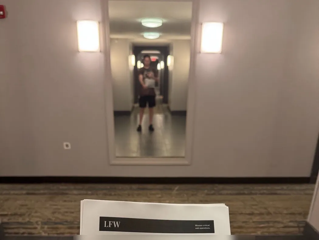 Leland holding up a printed LFW proposal, seen from behind the pages, with his reflection in a hallway mirror.