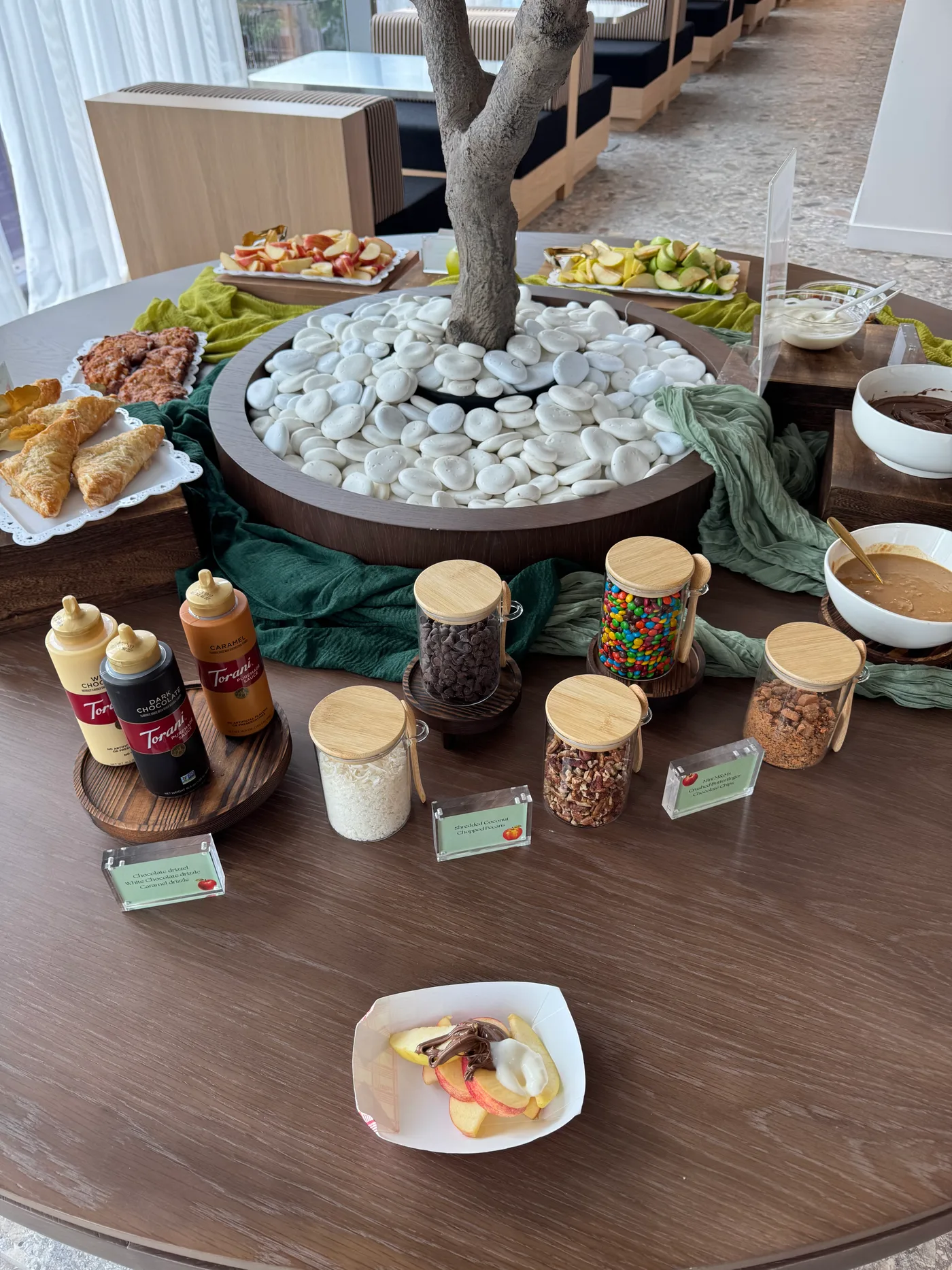A round table set with sliced red and green apples, jars of chocolate chips, candy and nuts, and bottles of chocolate and caramel sauce, around a tree planter in the WeWork Rosslyn lounge.