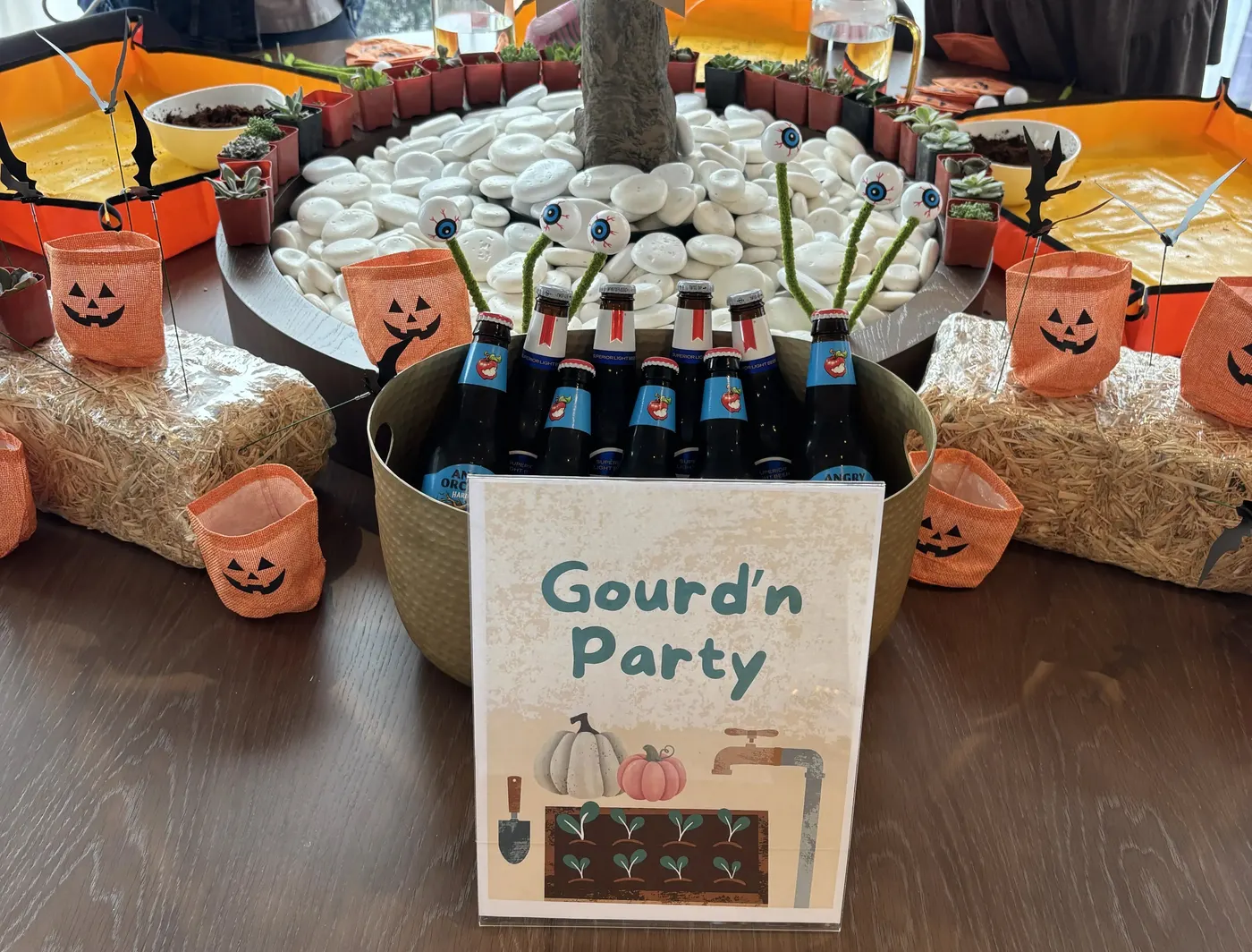 Un tavolo al WeWork Rosslyn allestito per un laboratorio di piantagione autunnale: un cartello con la scritta Gourd'n Party davanti a un cesto di bottiglie con scovolini a occhi mobili, piccole piante grasse in vaso intorno a una fioriera con un albero e sassi bianchi, balle di fieno e sacchetti arancioni a forma di zucca.