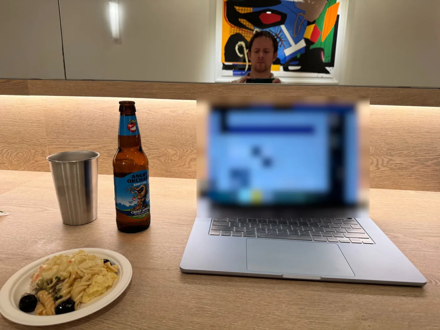 Leland a un bancone del WeWork Rosslyn, il viso riflesso nello specchio sotto un quadro astratto colorato, con davanti un piatto di insalate di pasta e di patate, un bicchiere di acciaio, una bottiglia di sidro Angry Orchard e un portatile. Lo schermo del portatile è sfocato.