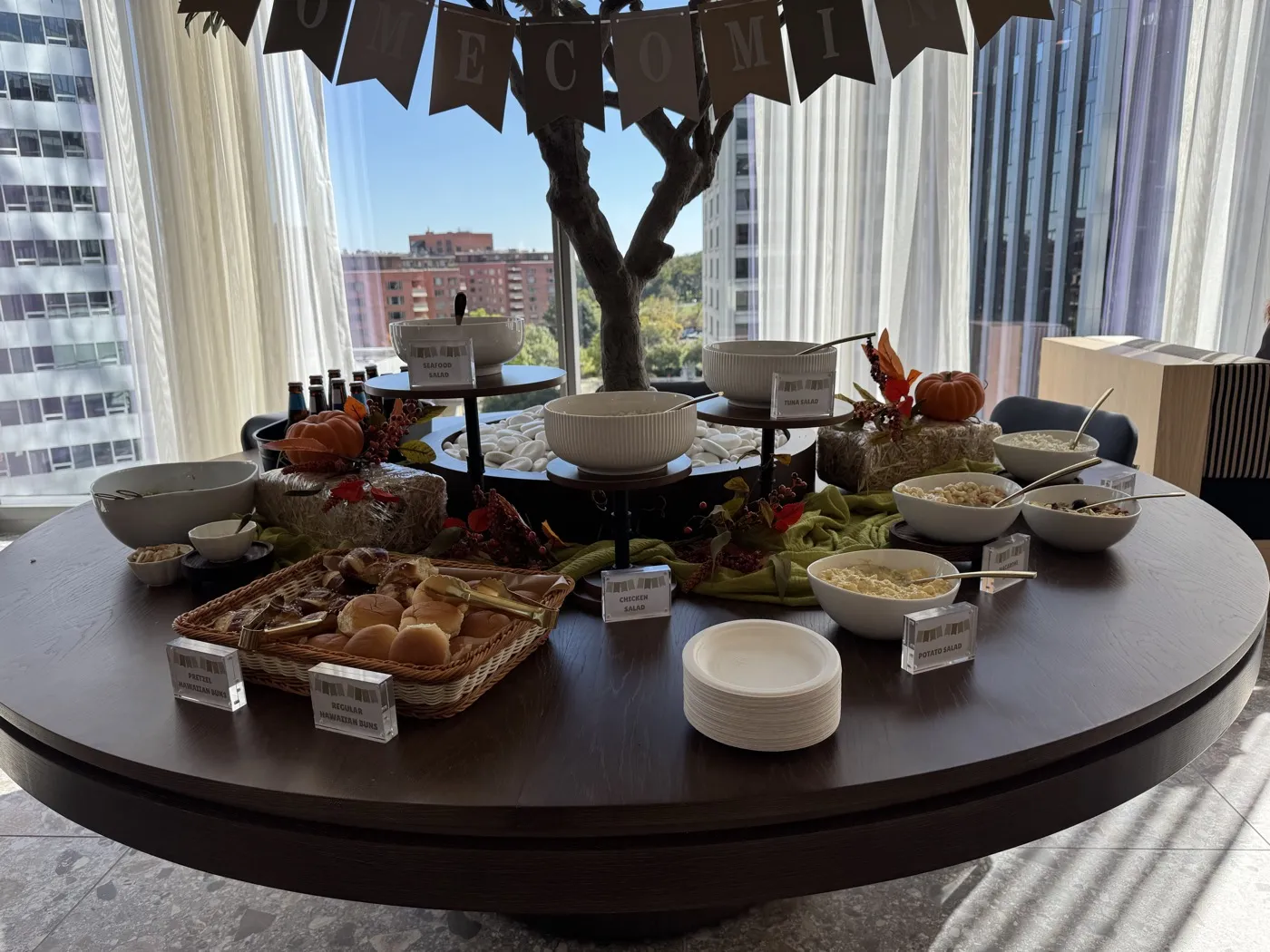 Un tavolo rotondo scuro al WeWork Rosslyn sotto una ghirlanda di bandierine che compone la parola HOMECOMING: insalatiere con i cartellini, un cestino di panini, piccole zucche e una pila di piatti di carta, con edifici per uffici oltre le finestre.