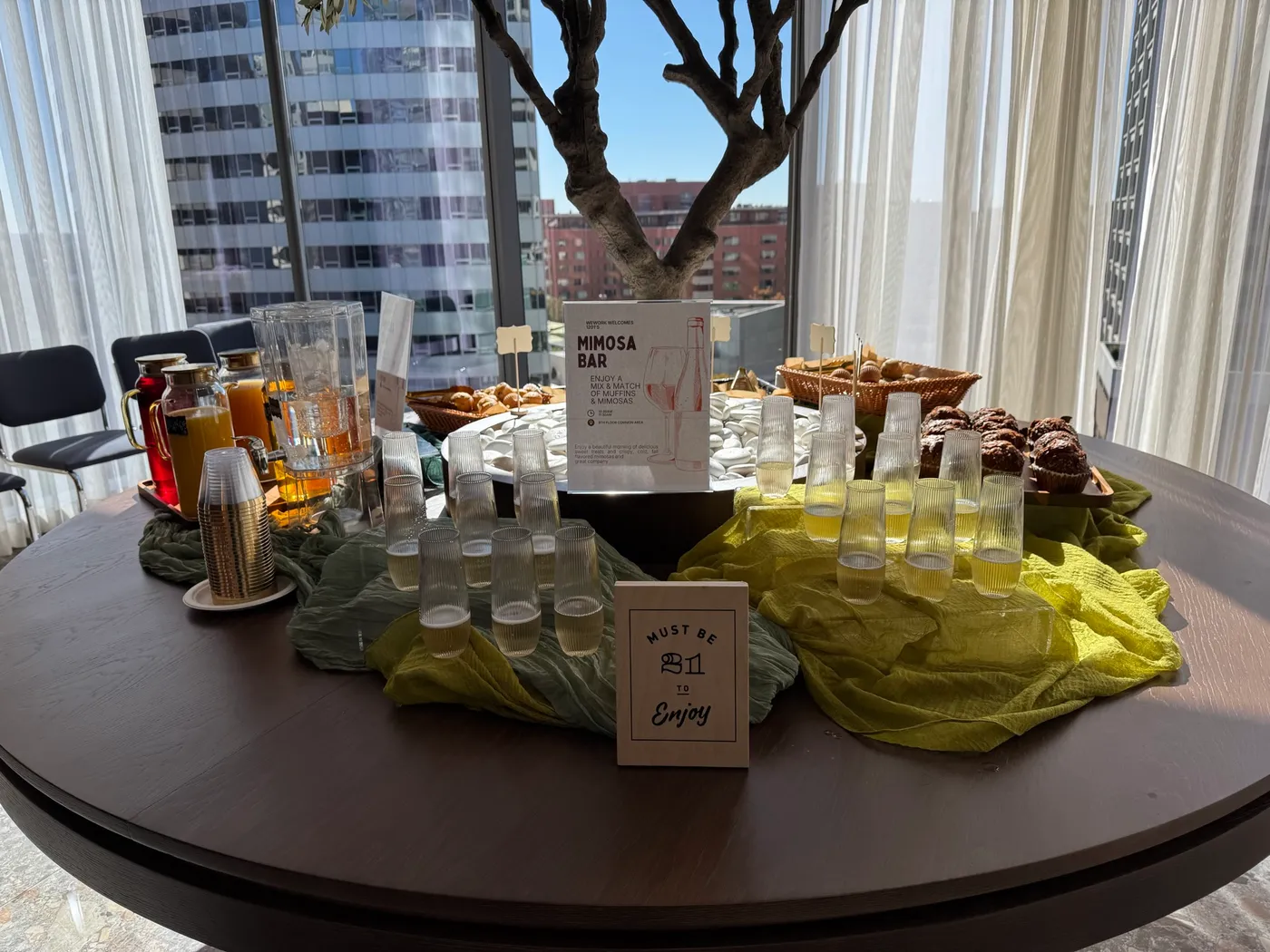 Il tavolo del Mimosa Bar al WeWork Rosslyn: un cartello che dà il benvenuto al Mimosa Bar del 1201, con muffin e mimosa dalle 10:30 alle 11:30 nell'area comune del nono piano; bicchieri di mimosa su teli verdi e gialli, un cartello Must be 21 to enjoy, caraffe di succo e cestini di muffin, con le torri di Rosslyn oltre la finestra.