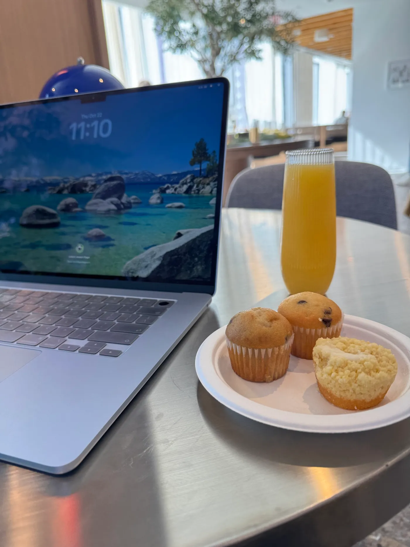 Un MacBook su un tavolo con la schermata di blocco alle 11:10 di giovedì 22 ottobre, accanto a un bicchiere di mimosa e un piatto con tre mini muffin.
