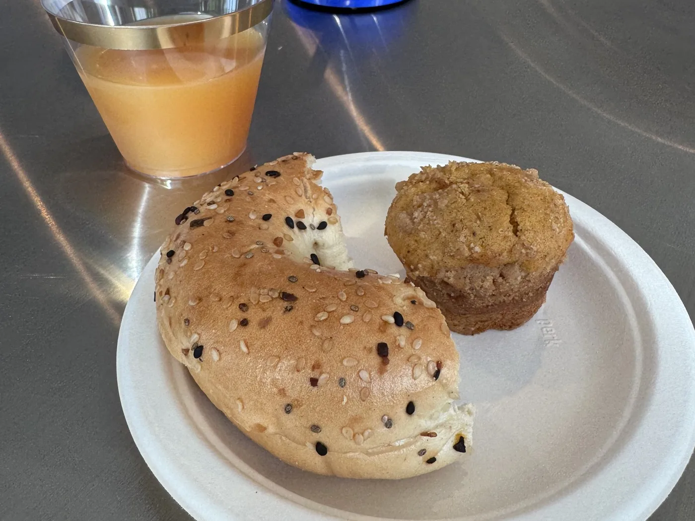 A paper plate with an everything bagel and a pumpkin muffin beside a cup of cider on a steel table at WeWork Rosslyn.