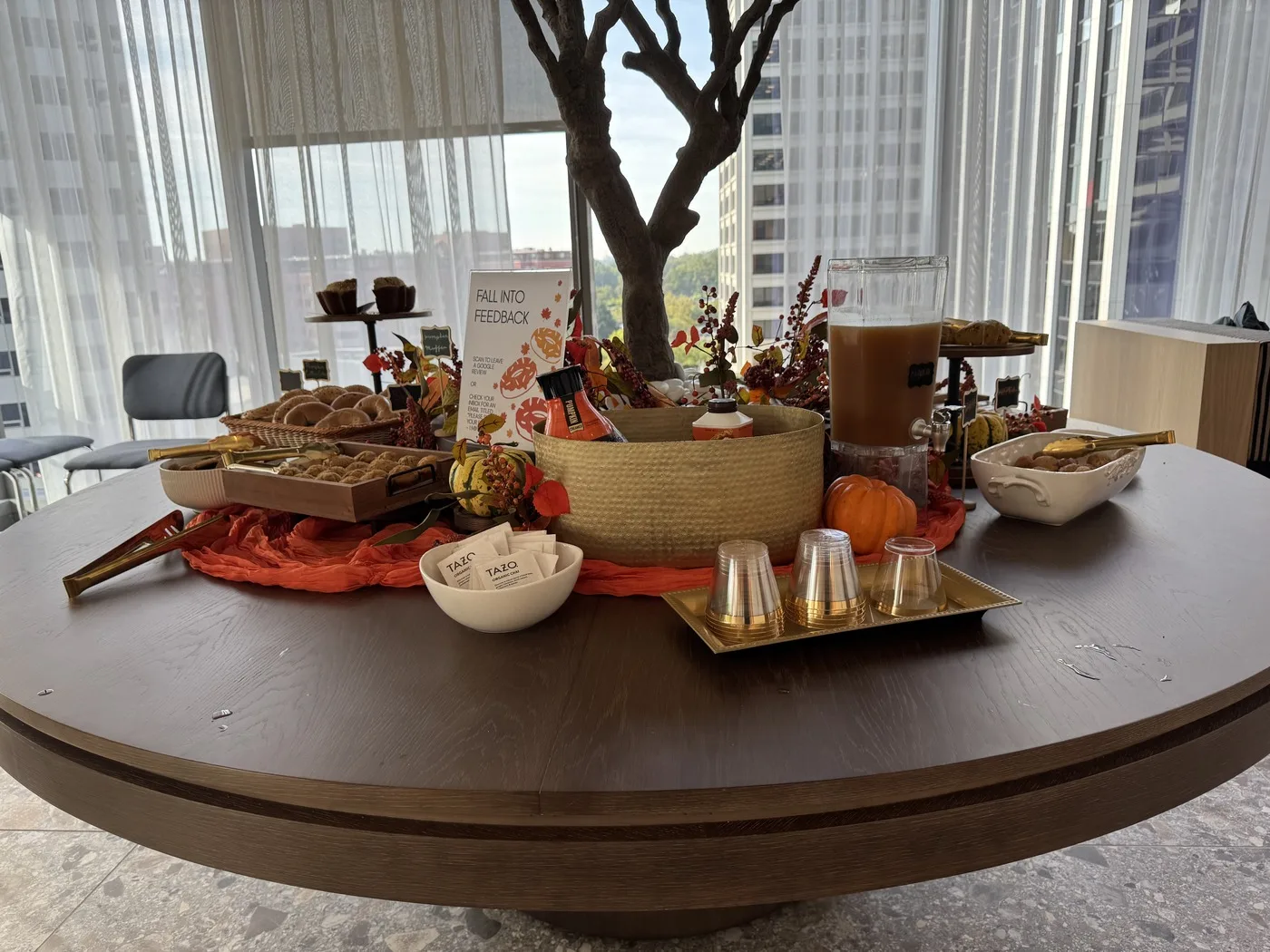 A round wooden table at WeWork Rosslyn dressed for fall: an orange runner, small pumpkins, a dispenser of cider, trays of bagels and muffins, Tazo tea bags, and a sign reading Fall Into Feedback, with office towers through the window behind.