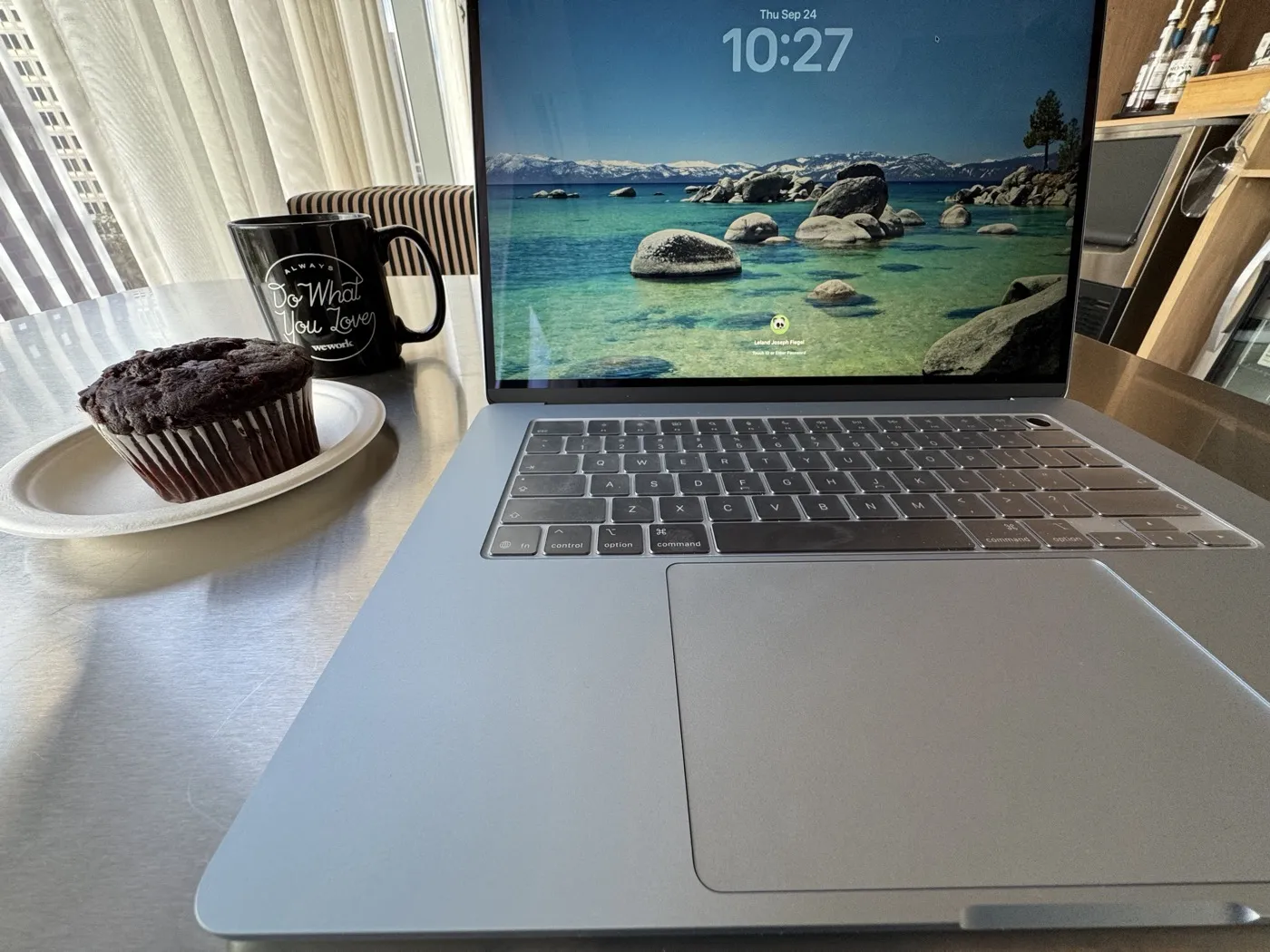 A chocolate muffin on a paper plate beside a black WeWork mug reading Always Do What You Love, next to an open laptop on a common-area table.