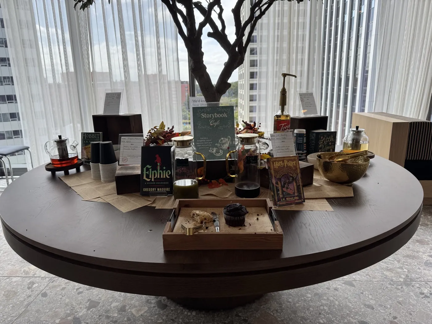 A round table at WeWork Rosslyn set up as a Storybook Cafe: a Storybook Cafe sign, jars of matcha and cold brew, a teapot, Elphie, The Hobbit and Harry Potter books standing among the drinks, and a wooden tray with a muffin and a scone.